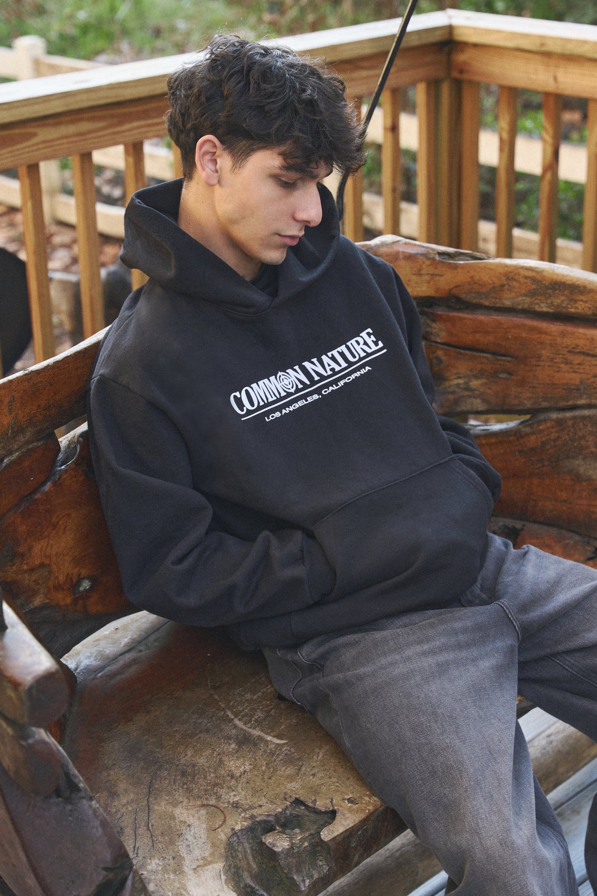 Man in a black Common Nature hoodie seated on a wooden bench outdoors