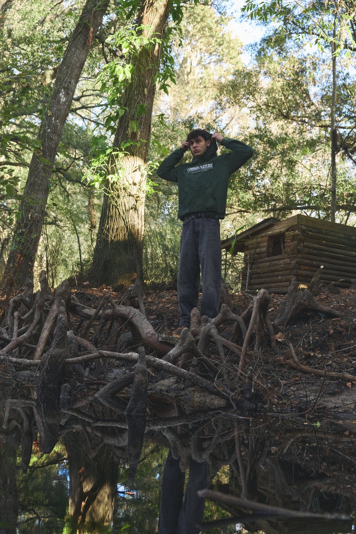 Man in a forest green Common Nature hoodie standing on fallen branches in the woods