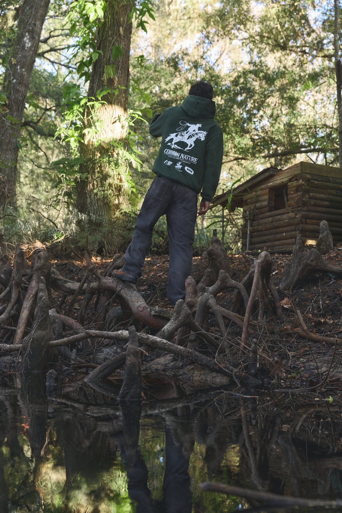 Back graphic of a forest green Common Nature hoodie in the woods beside a small cabin