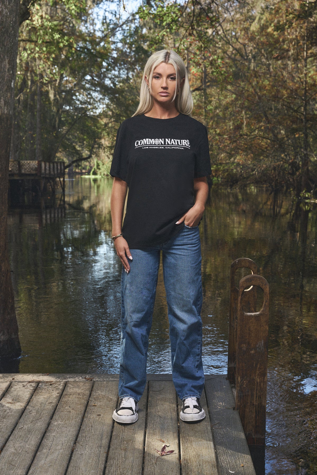Woman in a black Common Nature tee and jeans on a dock by the water