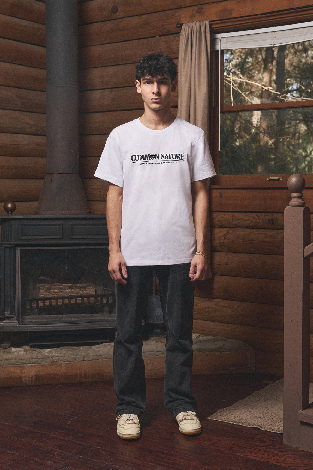 Man in a white Common Nature tee by the wood stove inside the cabin
