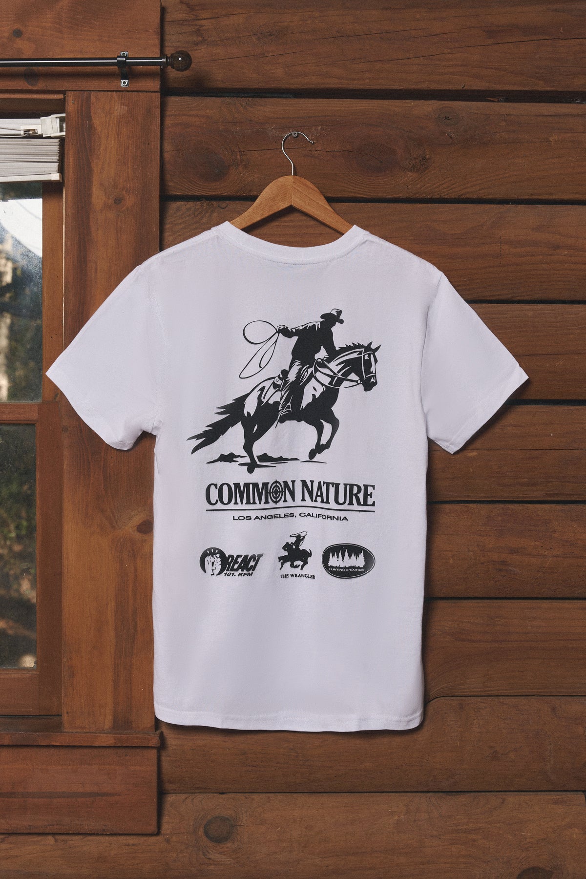 White Common Nature graphic tee hanging on a cabin wall
