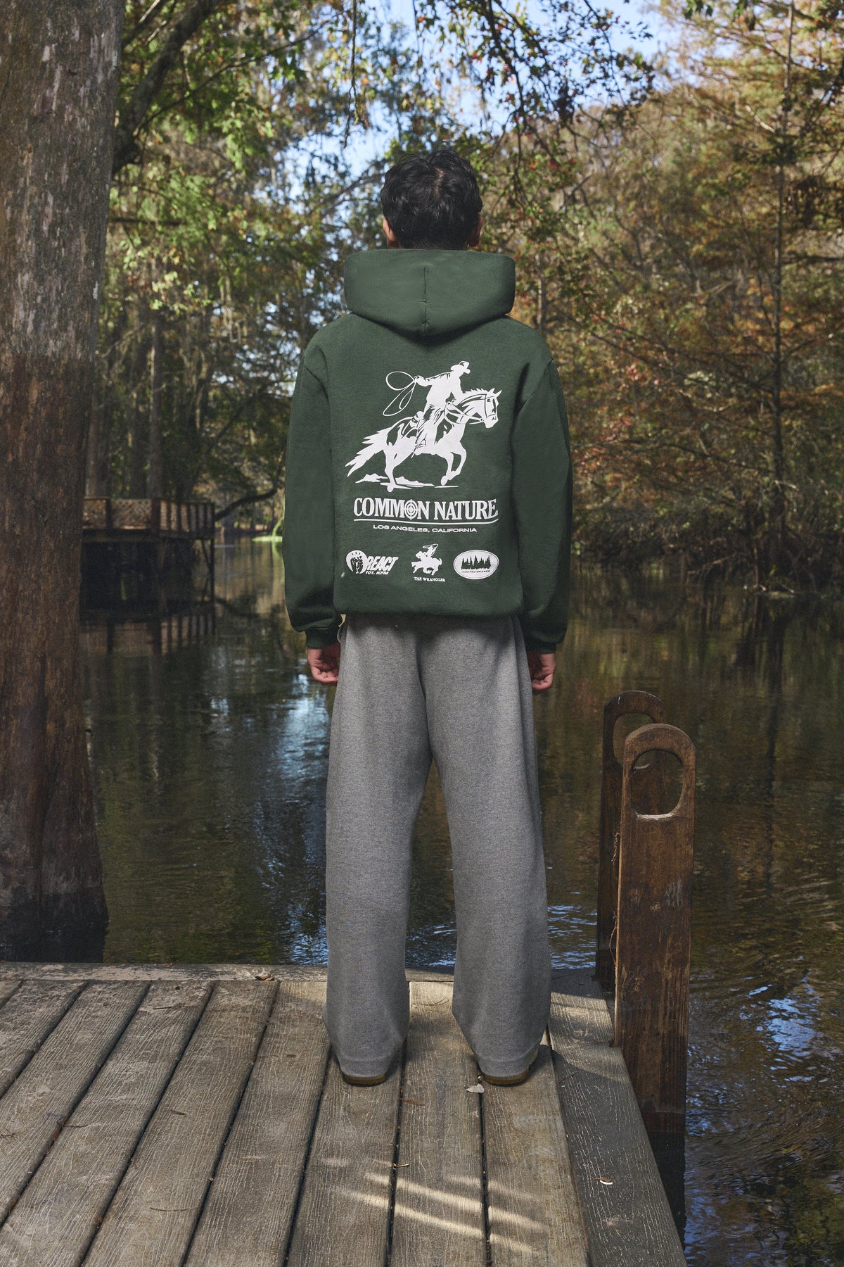 Back graphic of a forest green Common Nature hoodie on a dock by the water