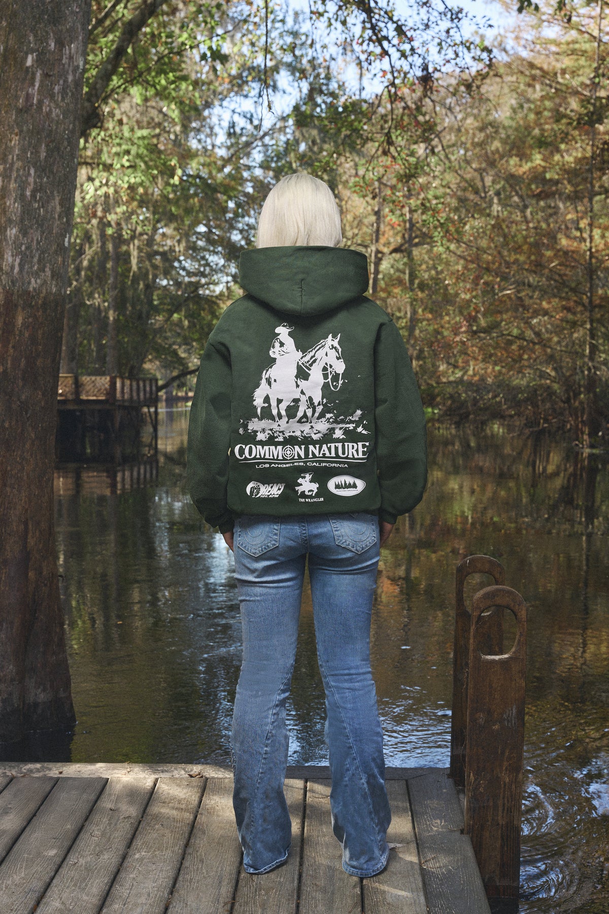 Back graphic of a forest green Common Nature hoodie on a woman by the water