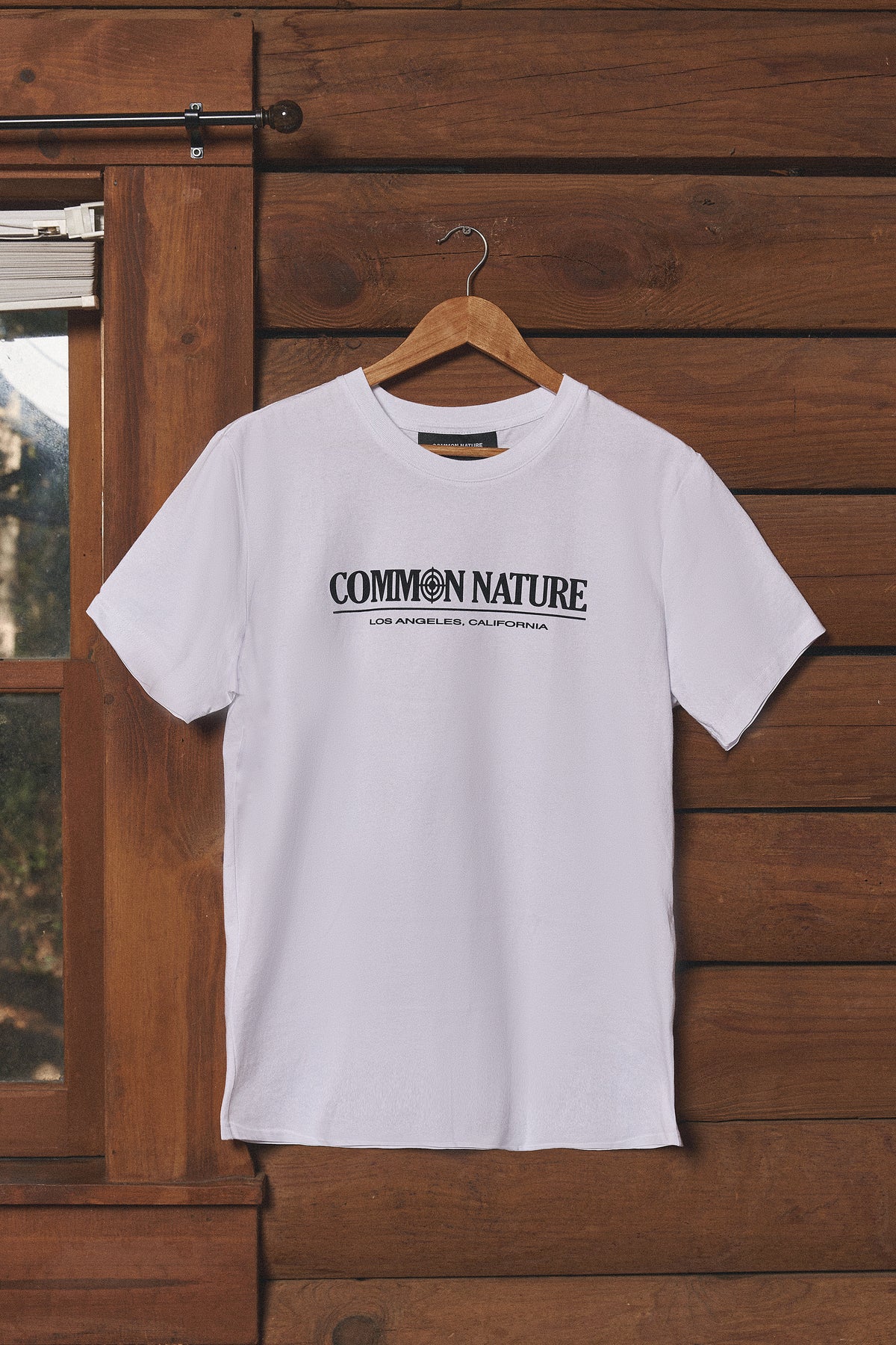 White Common Nature tee hanging on a cabin wall, front view