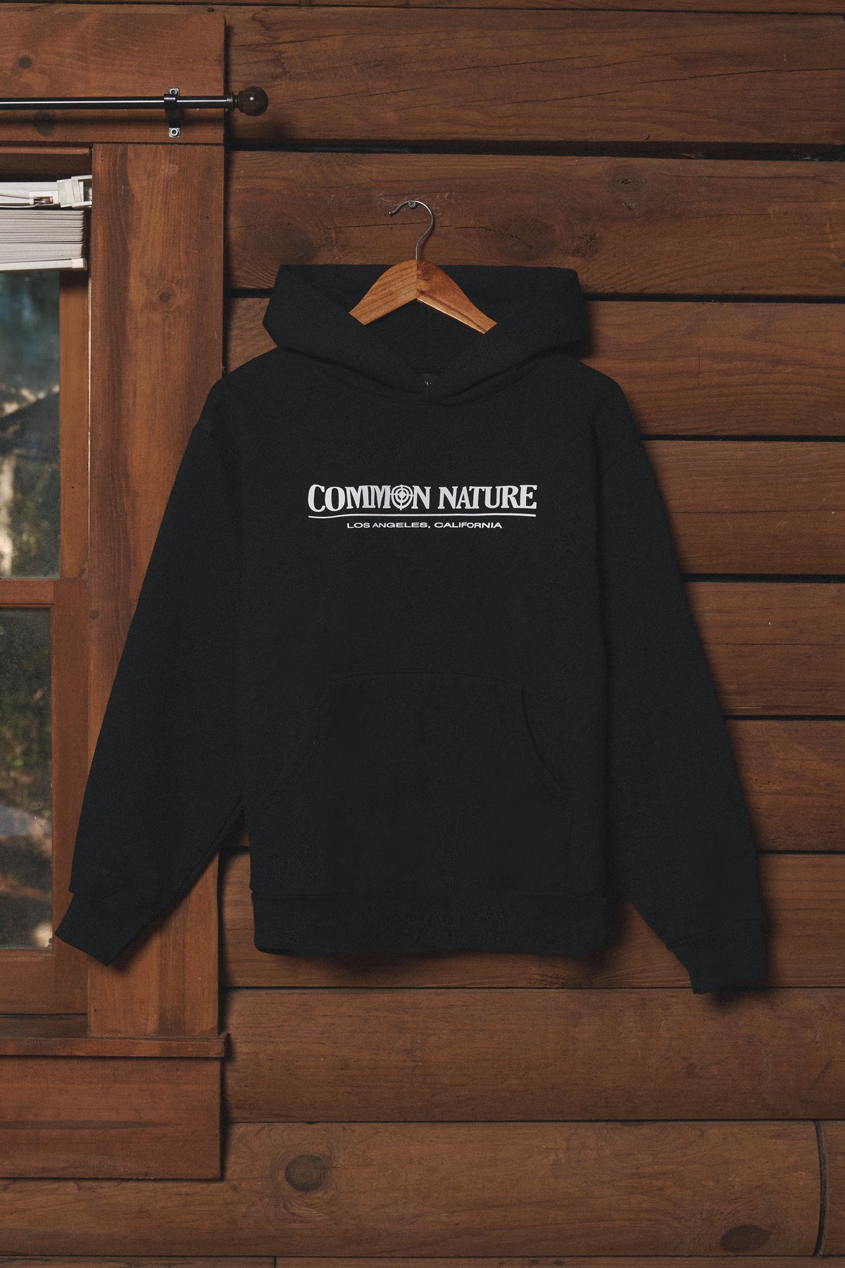 Black Common Nature hoodie hanging on a cabin wall, front view